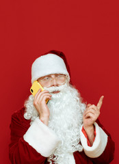 Santa Claus talking on the phone on a red background and looking away. Christmas greeting on the phone from Santa. Isolated. New Year concept. Vertical studio photo. Copyspace