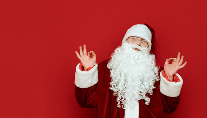 Portrait of satisfied Santa Claus with closed eyes shows hands gesture Ok isolated on red background. Copyspace on the right. Portrait of happy santa meditating on red background. X mas