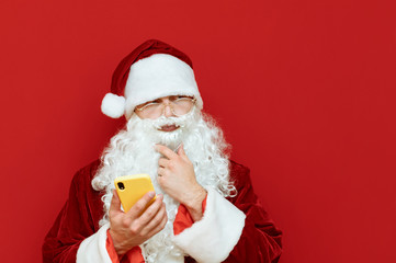 Studio photo of funny Santa with smartphone in hand on red background, looking away at blank space with pensive face. Santa with Phone and Copyspace. X-mas concept