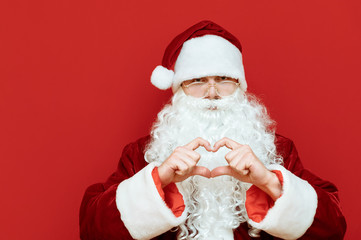 Closeup portrait of cute Santa Claus on red background, shows heart gesture and looks into camera. Santa loves Christmas. New Year concept. Copyspace X-mas