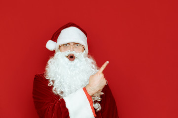 Closeup portrait of shocked Santa Claus isolated on red background, pointing sideways at copy space and looking surprised at camera with astonished face. Santa points to a blank space for text.