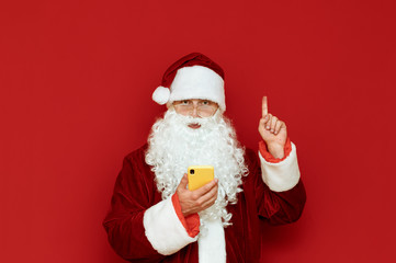 Fototapeta na wymiar Portrait of a funny Santa Claus standing on a red background with a smartphone in his hand, looking into the camera and pointing his finger up at an empty space copyspace. X-mas