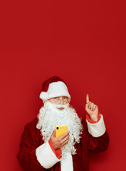 Santa Claus stands on red background with smartphone in hand, looks into camera with serious face and shows thumb up on copyspace. Isolated portrait. Vertical photo.