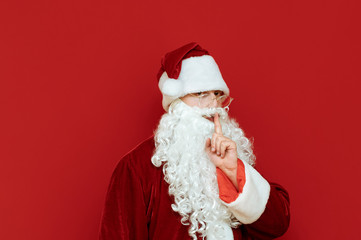 Serious Santa showing a gesture of silence on a red background, looking into the camera with a serious face. Tsh. Santa asks for silence, isolated. Christmas concept. Copyspace