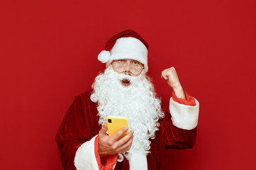 Portrait of joyful Santa Claus with smartphone in his hand rejoices with raised hand, shouts and looks into camera, isolated on red background. Happy Santa rejoices in victory. Xmas concept