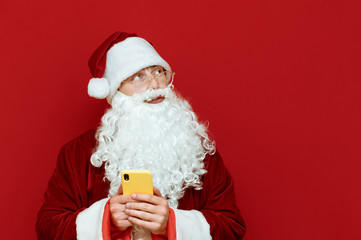 Portrait of pensive santa claus standing on red background with smartphone in hand and looking away at blank copyspace. Thoughtfully Santa isolated on red. Christmas and New Year concept.