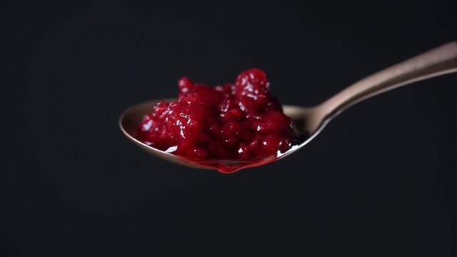 Spoon with red raspberry jam and one drop. Red raspberry jam dripping and flowing down from the metal spoon on black background, close up. Brass spoon with homemade red raspberry jam