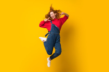 Redhead woman with overalls jumping over isolated yellow wall