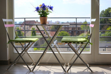 Two metal chairs with green pillows stand near the window. Chairs are at the table near the balcony. A bouquet of irises on the table. Garden furniture