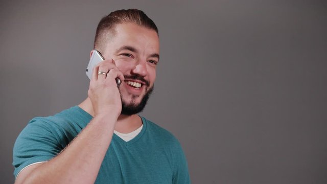 Young man with dark hair and beard, blue t-shirt holding smartphone over gray isolated background vigorously talks, with relish and smile