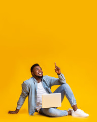 Black guy sitting with laptop and pointing at copy space