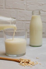 Oat milk pouring into a glass cup with a bottle of milk on the background, selective focus
