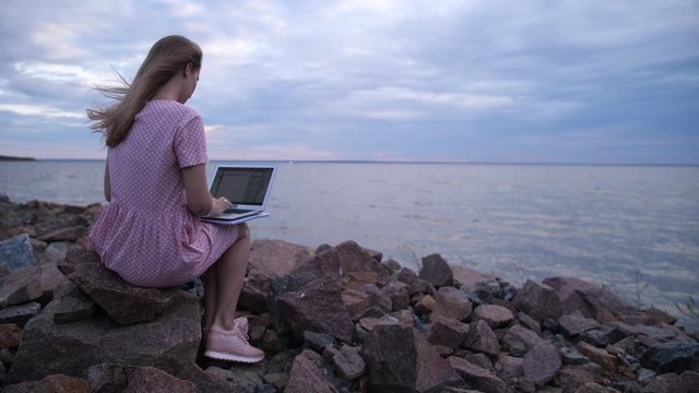 The blonde is sitting on the stones by the sea. She is typing on a laptop. Looking for inspiration. 4K Slow Mo