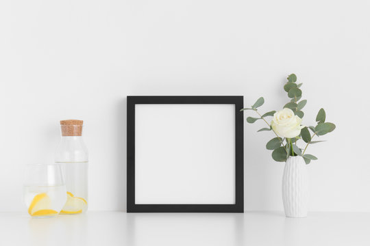 Black square frame mockup with a rose and eucalyptus in a vase, glass and a bottle on a white table.
