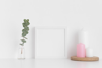 White square frame mockup with candles and eucalyptus in a vase on a white table.