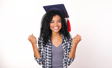 Finally! Graduate African American girl in a plaid shirt is standing and holding both of her palms in fists and smiling with her eyes closed, expressing success or victory.