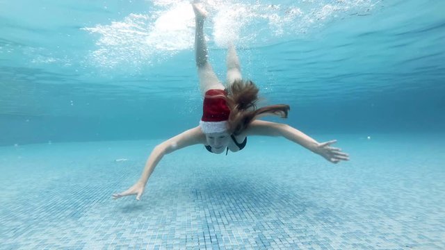 A young sexy girl in a red Santa hat dives and swims near the bottom of the pool on a Sunny day. She looks and poses for the camera with her hair down and waves her hands. Portrait. Slow motion. 4K.