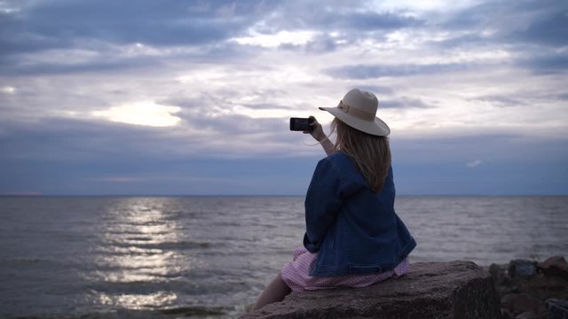 A young woman sits on a stone and takes a photo on the phone. The girl photographs the sunset sky. 4K Slow Mo