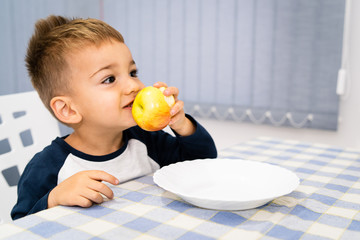 Portrait of small little boy three years old eating an apple fruit by the table at home