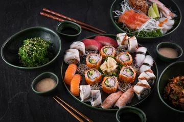Fotobehang Geassorteerde sushi set geserveerd op donkere stenen leisteen achtergrond. Zeewier wakame salade, zeevruchten, diverse maki rolls, sashimi en nigiri met kaviaar, garnaal, coquille, octopus, zalm, paling, makreel en tonijn © CrunchyBeans