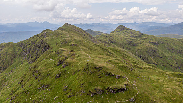 An aerial view of a grassy mountain rocky summit and mountain range in the background under a cloudy sky