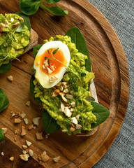Close up of an avocado toasts with soft boiled egg, herbs, spinach, salad and nuts on a round wooden board on grey background. Healthy food concept. Healthy breakfast