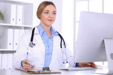 Young woman doctor at work in hospital looking at desktop pc monitor. Physician controls medication history records and exam results. Medicine and healthcare concept