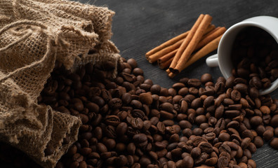Grains of coffee pour out from a white coffee cup on a dark background from a bag with cinnamon sticks