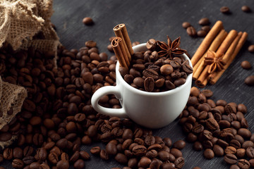 White cup full of coffee beans cinnamon bag spills out coffee stars anise star anise on a dark background
