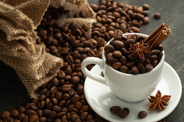 White cup full of coffee beans cinnamon bag spills out coffee stars anise star anise on a dark background