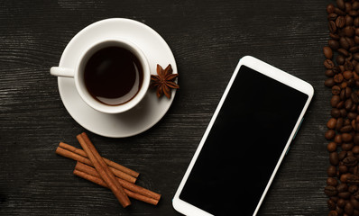 White cup with cinnamon and anise coffee sticks and a mobile phone with a blank screen on a black background with coffee beans and place for text top view
