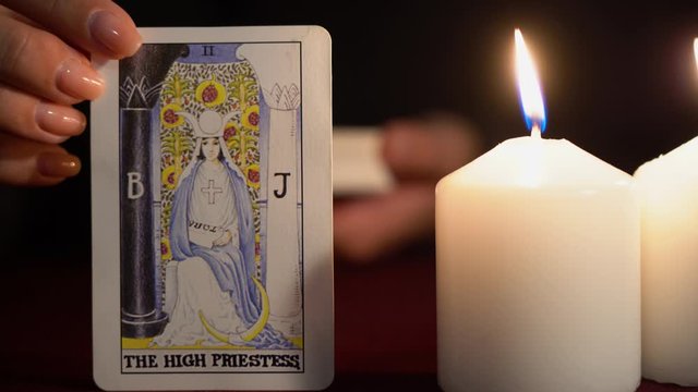 A witch is fortune teller in black mantle with manicure shows tarot card of 2 Arcana: The High Priestess. White candles burns on table. Occult, esoteric, divination and wicca concept. Magician guesses