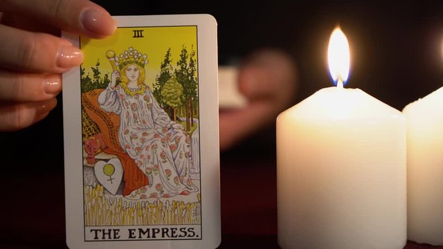 A witch is fortune teller in black mantle with manicure shows tarot card of 3 Arcana: The Empress. White candles burns on table. Occult, esoteric, divination and wicca concept. Magician guesses.