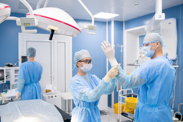 Young assistant putting gloves on hands of surgeon while preparing for operation
