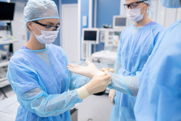 Young nurse in protective mask and workwear helping surgeon with gloves