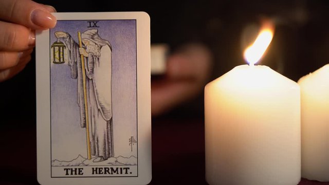 A witch is fortune teller in black mantle with manicure shows tarot card of 9 Arcana: The Hermit. White candles burns on table. Occult, esoteric, divination and wicca concept. Magician guesses.