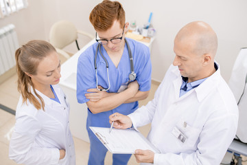 Bald serious doctor pointing at document while reading it aloud to young interns