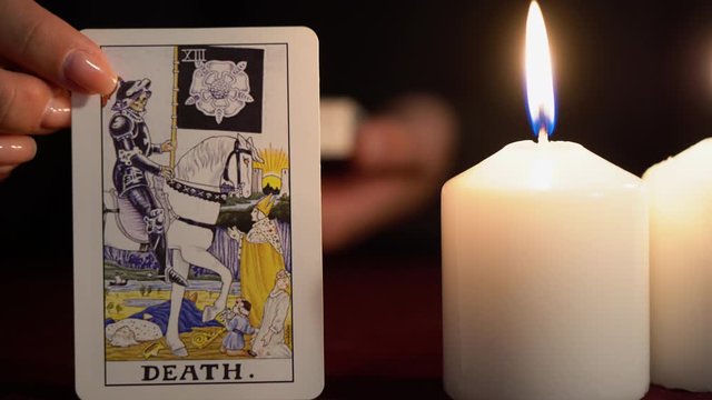 A witch is fortune teller in black mantle with manicure shows tarot card of 13 Arcana: Death. White candles burns on table. Occult, esoteric, divination and wicca concept. Magician guesses.