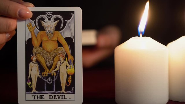 A witch is fortune teller in black mantle with manicure shows tarot card of 15 Arcana: The Devil. White candles burns on table. Occult, esoteric, divination and wicca concept. Magician guesses.