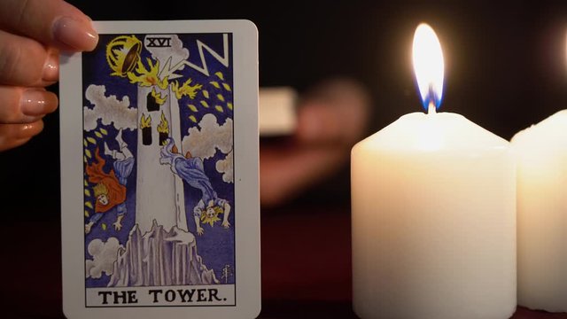 A witch is fortune teller in black mantle with manicure shows tarot card of 16 Arcana: The Tower. White candles burns on table. Occult, esoteric, divination and wicca concept. Magician guesses.