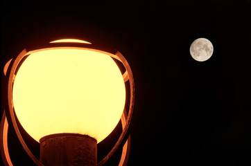 In the dark, a spherical street lamp made of frosted glass shines. In the background is a round moon with a distinguishable surface structure. Background.