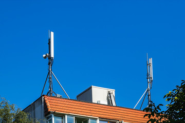 Two antennas of cellular communication on an orange tiled roof of a multi-storey residential building against blue sky.