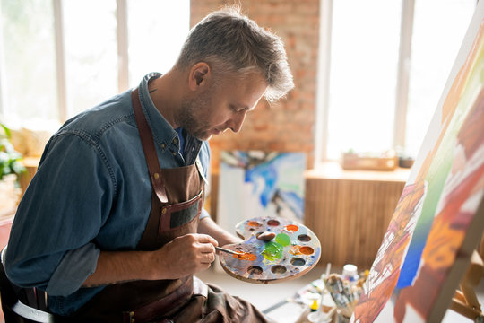 Creative man in apron mixing colors on palette in front of easel in studio