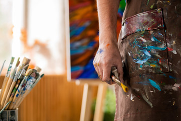 Hand of contemporary artist in apron holding paintbrush while working in studio