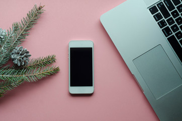 top view desktop, computer, phone with Christmas decorations, mobile greetings, New Year holidays, gifts, year planning, spruce branch, pine cone, snow on pink background. flat lay; copy space; text.