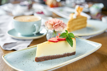 Piece of delicious cheesecake with strawberry and mint leaves on white plate. Breakfast in the cafe, morning coffee. Cappuccino and lots of desserts on the table.
