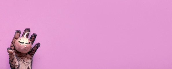 Hand in black mesh glove holding pink pumpkin with closed eyes. Creative Halloween panoramic flat lay on pink paper background with copy-space