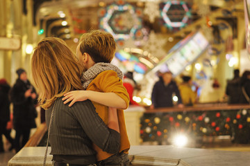 Dressed in stylish modern casual wear, mother and son in shopping mall decorated for Christmas. Garlands and christmas lights on background in defocus. Back view. Lifestyle concept. Copy space