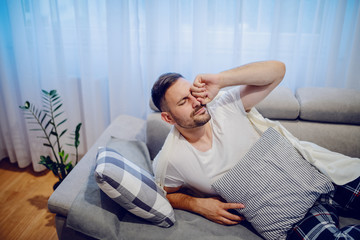 Handsome sick caucasian unshaven man in pajamas waking up after afternoon nap. Living room interior.