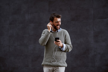 Handsome caucasian fashionable man in sweater putting earphones and holding smart phone while standing outside in front of gray wall.